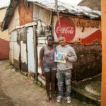 Two people standing outside a rustic shack.