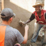 Two men plastering a wall together, smiling.